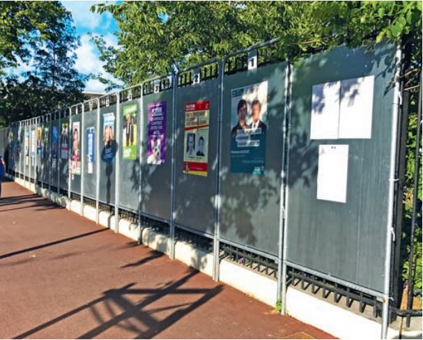 photo de plusieurs panneaux électoraux avec des affiches de candidats collés dessus installés dans une rue sous les arbres